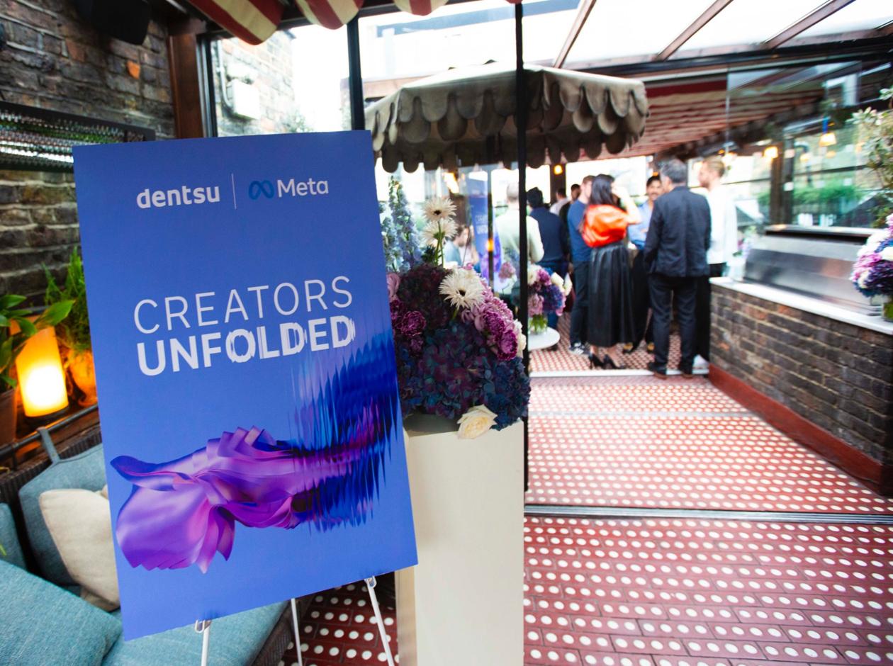 A large blue poster with the dentsu and Meta logos reading “Creators Unfolded” displayed at an indoor event with people mingling in the background and floral decorations beside it.